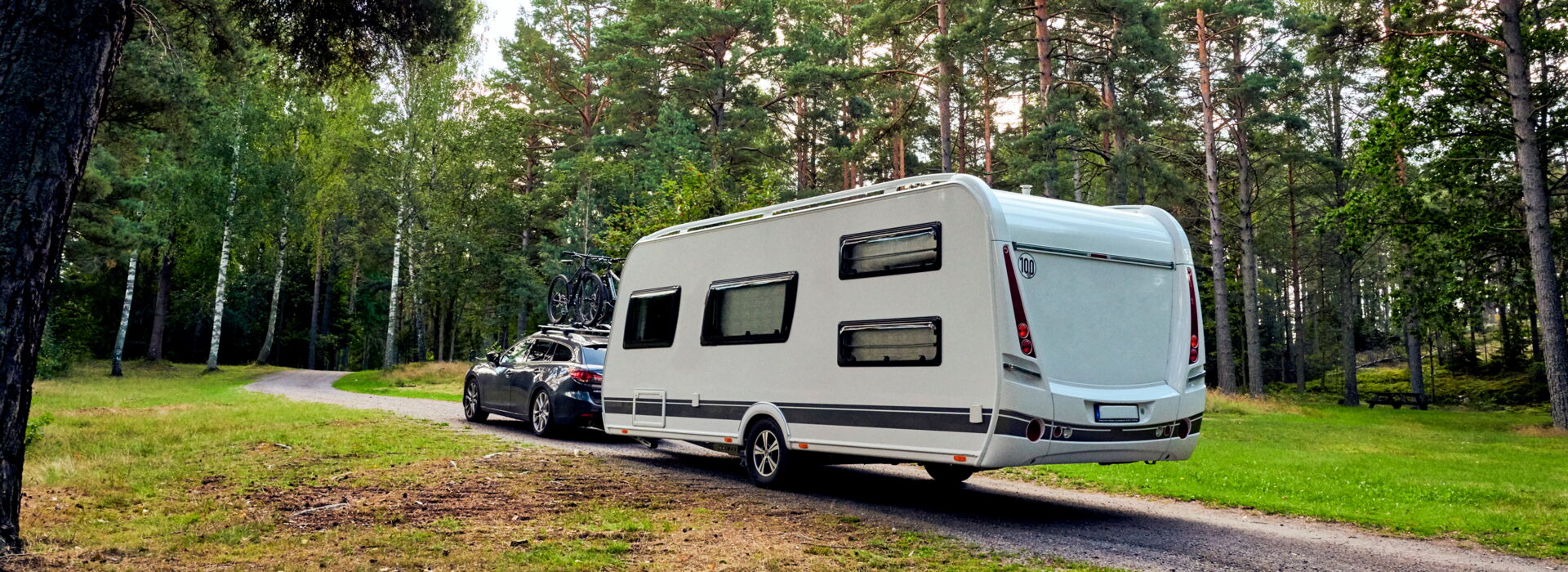 Ein Auto zieht einen weißen Dethleffs Nomad Wohnwagen auf einer Waldstraße. Reifendienst Böhme in Vetschau bietet Reifenservice für Ihre Reise.