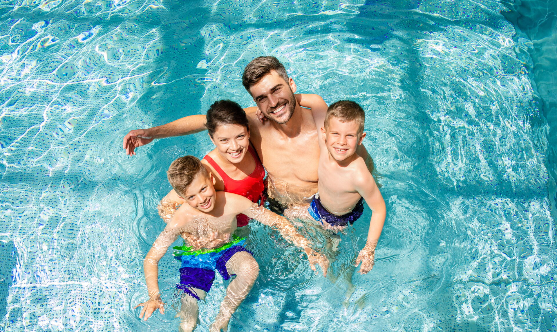 Freizeitspaß für die Familie | Reifendienst Böhme Vetschau Eine glückliche Familie mit zwei Kindern schwimmt in einem hellblauen Pool. Reifendienst Böhme in Vetschau wünscht Ihnen eine schöne Freizeit.